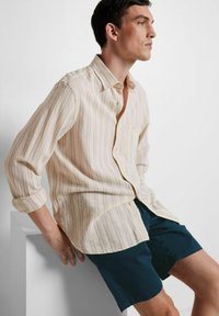 Camicia a maniche lunghe a righe chiare con colletto a punta e taschino sul petto, abbinata a pantaloni corti blu scuro. Il tessuto sembra essere strutturato.