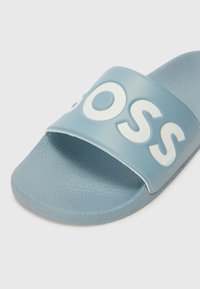 Blå skyvesandal laget av teksturert gummi. Har et bredt, matt stropp med hvite hevede bokstaver som staver "BOSS." Flat, polstret såle.