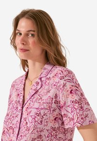 Korte mouwen pyjama top in roze stof, met een bloemenpatroon in donkerdere rozen en paars, met een borstzak en contrasterende afwerking.