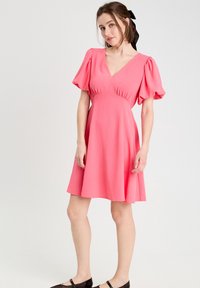 Robe rose à manches courtes avec un décolleté en V, un corsage froncé et une jupe évasée, réalisée en tissu léger. Des chaussures plates noires complètent la tenue.
