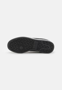Black rubber shoe sole with a textured surface. Features a circular emblem and raised tread pattern for grip. A narrow white stripe runs along the side.