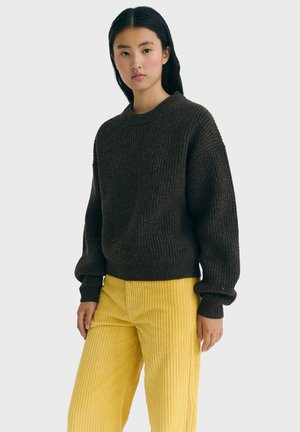 Junge Frau in einem dunkelbraunen Rippenpullover und leuchtend gelben Cordhosen, die vor einem einfachen hellen Hintergrund steht.