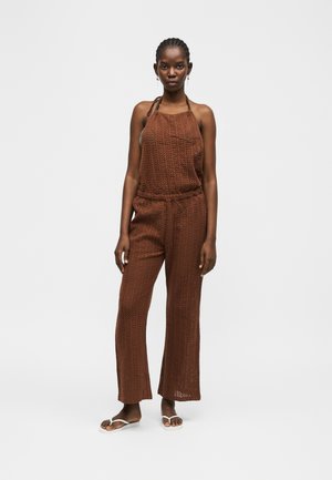 Młoda kobieta ubrana w brązowy, szydełkowy top na szyję bez rękawów oraz pasujące szerokie spodnie i białe klapki, stojąca na tle jednolitym.