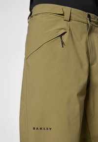 Pantalon imperméable vert avec une texture lisse, doté d'une poche zippée et d'une taille ajustable. Comprend un marquage noir "OAKLEY".