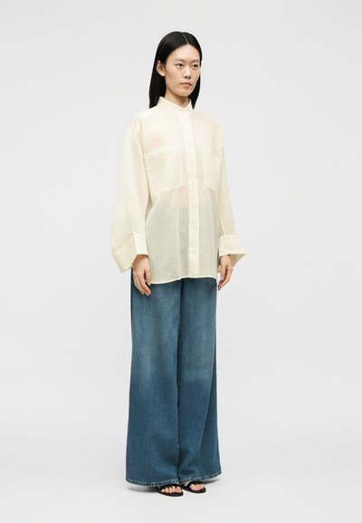 Femme debout portant une chemise crème transparente boutonnée avec de larges manches, un jean bleu à jambes larges et des sandales noires sur un fond uni.