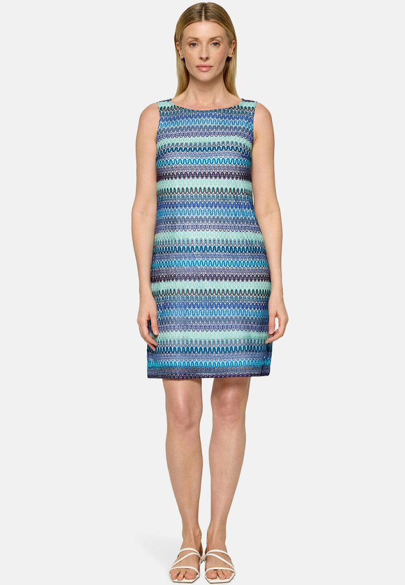 Femme debout portant une robe sans manches, longueur genou, avec des rayures ondulées horizontales bleues, turquoise et noires, et des sandales blanches à bouts ouverts.