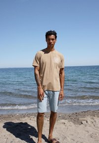 Άνδρας που στέκεται σε αμμώδη παραλία, φοράει μπεζ μπλουζάκι, ανοιχτόχρωμα τζιν σορτς και καφέ σανδάλια, με φόντο τον ωκεανό και καθαρό ουρανό.