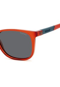 Lunettes de soleil transparentes rouges avec des verres gris, forme rectangulaire et logo Polaroid sur les accents bleus des branches. Texture en plastique lisse.