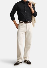 Homme portant une chemise noire boutonnée, un pantalon blanc avec une ceinture à motifs, des baskets foncées, tenant une veste beige sur l’épaule.