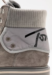 Primer plano de una bota de ante gris con un cuello acolchado y acanalado y un puño de nailon doblado, que presenta un logo negro y ojales de metal.