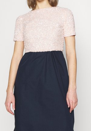 Kobieta ubrana w sukienkę z krótkim rękawem, jasnoróżową górą z cekinów i ciemnogranatową spódnicą, stojąca na tle jednolitego tła.