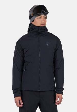 Black insulated jacket with a high collar and zip closure. Features a logo on the chest, and paired with a gray beanie and black goggles.