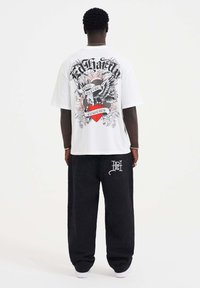 White short-sleeve t-shirt featuring large black and red graphic design on the back, paired with black pants and white sneakers.