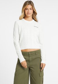 Supjaustytas baltas megztinis su kabeliniais raštais, su guminėmis rankovėmis ir apatine dalimi. Juodas „GUESS JEANS“ logotipas ant krūtinės. Suderintas su alyvuogių žalios spalvos kargo kelnėmis.