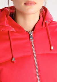 Czerwona pikowana kurtka z zamkiem błyskawicznym z przodu, kapturem ze sznurkami oraz srebrnymi elementami metalowymi. Gładka tekstura z przeszywanymi sekcjami.
