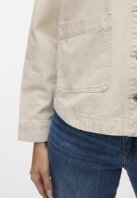 Beige bomullskappa med långa ärmar, knappstängning fram och två sidofickor. Bärs över mörkblå jeans med en ren, slät textur.