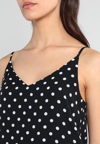 Svart ärmlös topp med V-ringning, prydd med ett mönster av vita prickar på ett jämnt tyg. Tunna axelremmar och lös passform.
