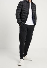 Man som bär en svart pufferjacka, vit t-shirt med rund hals, svarta joggingbyxor med vit text, och vita sneakers, står med händerna i fickorna.