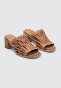 Mules de tacón abiertos en color tostado, con un diseño trenzado, una silueta cuadrada y una superficie texturizada. El tacón tiene forma de bloque y es robusto.
