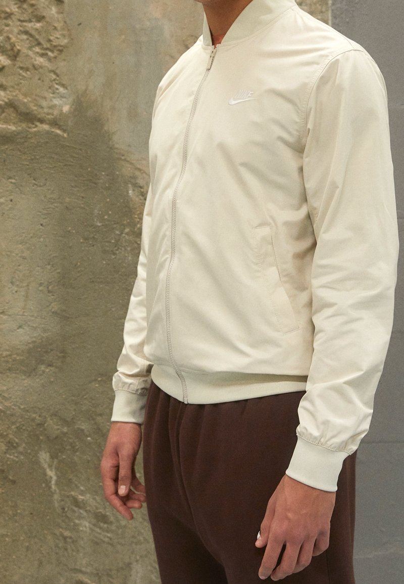 Homme portant une veste zippée Nike beige clair avec des poches et un pantalon marron, debout contre un mur texturé.