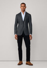 Dunkelgrauer Blazer mit Reverskragen, einer einzigen Knopfleiste und zwei frontalen Taschen, kombiniert mit einem hellblauen Hemd und schwarzen Hosen. Braune Wildlederschuhe.