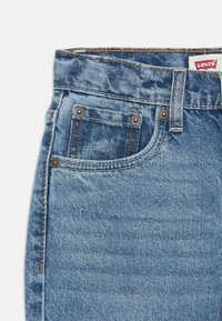Jeans de mezclilla de color azul claro con un diseño clásico de cinco bolsillos, costuras visibles, botones de metal y una etiqueta de marca en la cintura.