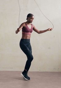 Frau in Sportbekleidung und Sneakers, die drinnen gegen eine schlichte Wand Seil springt.