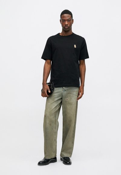 Hombre que lleva una camiseta negra con un pequeño zorro bordado, jeans verdes holgados, zapatos negros, sosteniendo un bolso negro contra un fondo blanco liso.