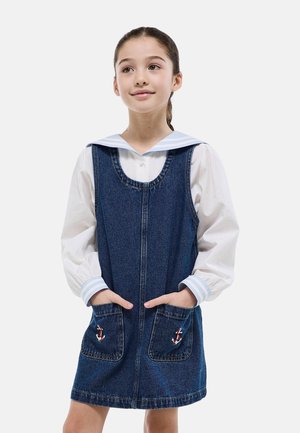 Fille aux cheveux bruns attachés en queue de cheval, portant un chemisier blanc avec col et poignets à rayures bleues sous une robe en jean avec des écussons d'ancre sur les poches.