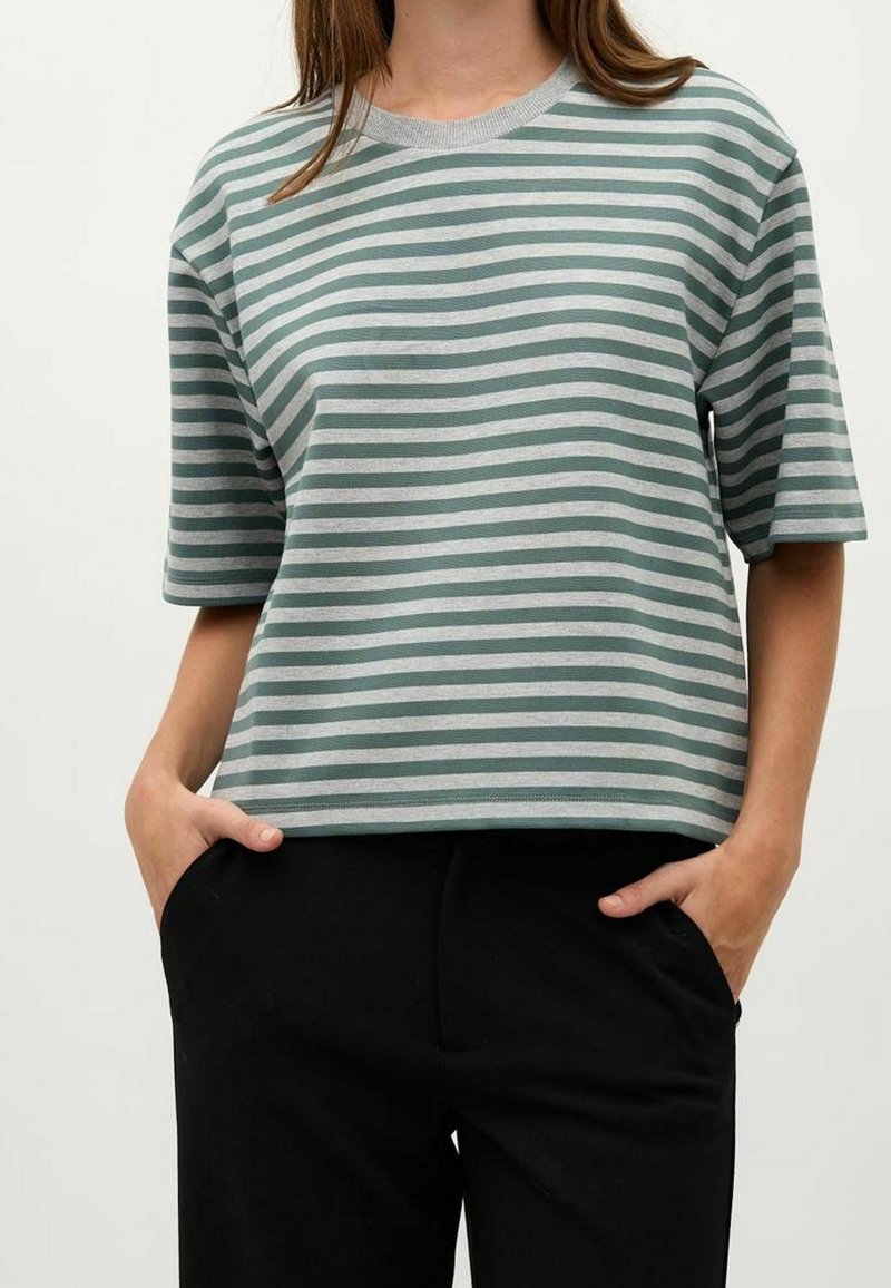 Personne portant un t-shirt ample à rayures horizontales vertes et grises à manches courtes avec les mains dans les poches d'un pantalon noir.