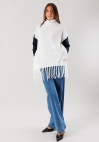 Maglione bianco oversize con colletto alto e orlo sfrangiato, realizzato in un materiale morbido, indossato sopra una maglia scura e abbinato a jeans blu e tacchi neri.