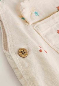 Tissu beige clair avec des motifs floraux en teal et orange. Présente un bouton doré avec "suivant" gravé, détail de poche partiellement visible.