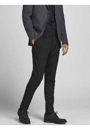 Homme portant un pantalon de costume noir, des chaussures noires, une chemise foncée et un blazer gris, debout, une main dans la poche, sur un fond uni.