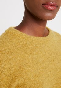 Suéter de punto texturizado amarillo mostaza con un cuello redondeado. El material suave y esponjoso resalta la apariencia acogedora de la prenda.