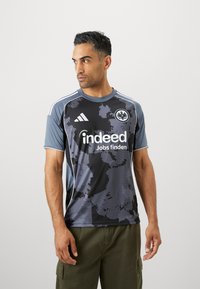 Jersey deportivo de manga corta en gris y negro con un patrón de formas irregulares. Tiene acentos en blanco y el logo de Adidas.