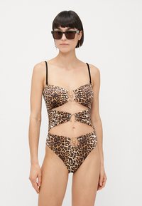 Costume da bagno intero a stampa leopardata con design a taglio, accenti dorati e sottili spalline nere. Il materiale appare liscio e texturizzato.