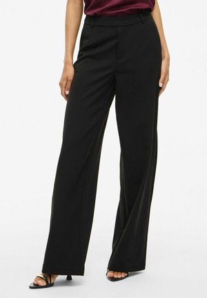 Pantaloni neri a gamba larga realizzati in tessuto liscio. Presentano una vestibilità sartoriale, una piega sottile e tasche laterali. Abbinati a sandali neri con cinturini.