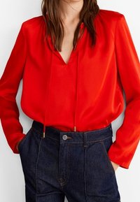 Blusa de satén roja con escote en V y detalle de cordón, combinada con vaqueros de mezclilla oscuros de tiro alto con cierre de botón metálico.