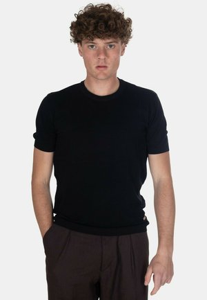 Giovane uomo con capelli ricci che indossa una maglietta nera semplice a maniche corte e pantaloni marrone scuro, in piedi con una mano in tasca.