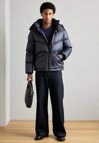 Jaqueta acolchoada preta com gola alta e fecho de correr, combinada com calças de ganga largas escuras e uma mala preta.