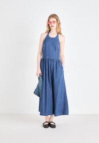 Robe maxi en denim avec un buste ajusté, une taille froncée, des poches latérales et une jupe fluide. Les accessoires comprennent des lunettes de soleil roses et une pochette en denim.