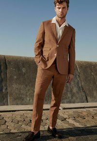Braun maßgeschneiderter Anzug mit Reverskragen und zwei vorderen Taschen. Darunter ein weißes Hemd mit Kragen. Schwarze Slipper runden das Outfit ab.