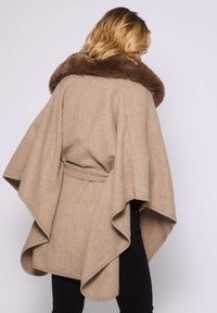 Femme avec des cheveux blonds portant une cape en laine beige avec un grand col en fourrure marron et un pantalon noir, vue de dos sur un fond blanc.