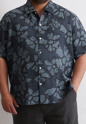 Mężczyzna w ciemnoniebieskiej koszuli z krótkim rękawem zapinanej na guziki, z dużymi jasnoniebieskimi kwiatowymi wzorami, ręka w kieszeni, widoczne ciemne spodnie.