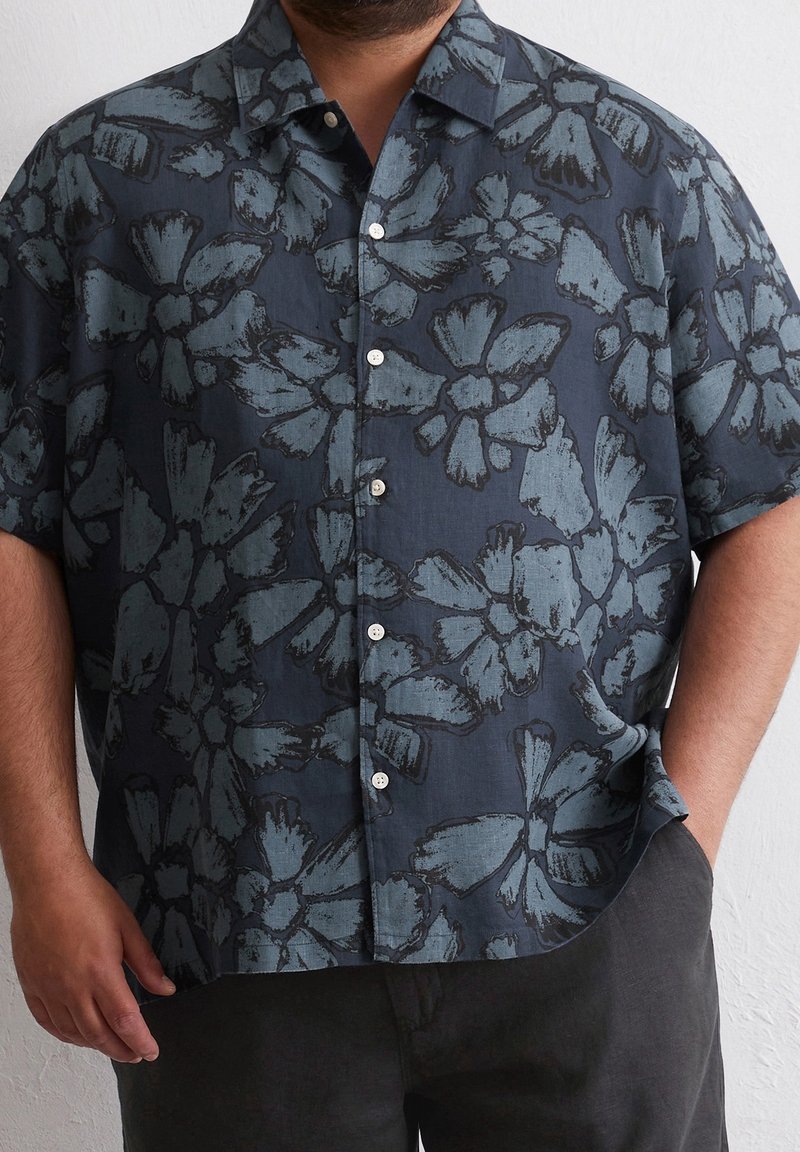 Homme portant une chemise à manches courtes bleu foncé à boutons avec de grands motifs floraux bleu clair, main dans la poche, pantalon foncé visible.