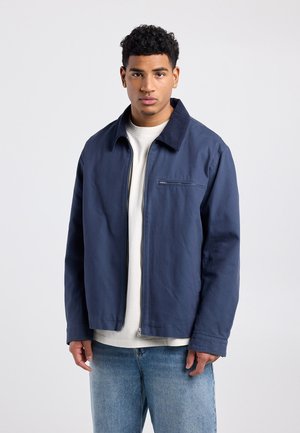 Young man wearing a navy blue zip-up jacket over a white shirt and light blue jeans, standing against a plain light background.