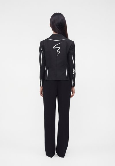 Γυναίκα με μακριά σκούρα μαλλιά που φοράει μαύρο σακάκι και παντελόνι, με την πλάτη προς τον θεατή, με λευκά αφηρημένα γραμμικά σχέδια στα μανίκια και την πλάτη του σακακιού.