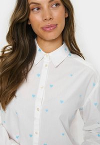 White button-up shirt with small blue heart patterns. Classic collar and placket with round, pearl-like buttons. Soft fabric texture.