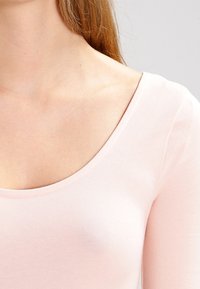 Světle růžový dlouhý rukáv s širokým výstřihem a hladkým, elastickým materiálem, který vykazuje vypasovaný střih a minimální švy.