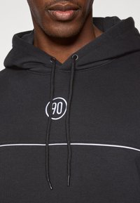 Melnā kapuce ar baltu izšūtu "90" logotipu, kontrastējoša balta svītra pāri krūtīm un regulējami auklas. Mīksta auduma tekstūra.